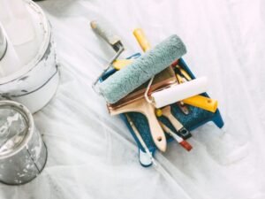A tray filled with paint rollers, brushes, and painting tools sits on a plastic-covered floor. Nearby are paint cans and a color swatch fan displaying various paint colors.