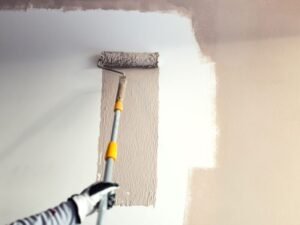 A hand in a glove uses a paint roller with an extension pole to apply beige paint to a white wall, partially covering the original color.