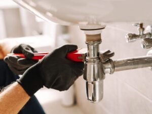 A person wearing black gloves uses a red pipe wrench to perform drain cleaning under a white sink.