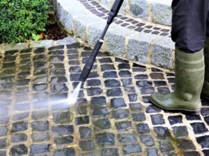 A person wearing green rubber boots power washes a stone-paved walkway, spraying water to clean the dark cobblestones near stone steps and a bush, demonstrating general handyman services offered by BrightWise Handyman Services.