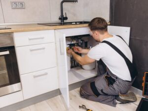 A plumber wearing safety goggles and overalls performs pipe repairs under a kitchen sink, with cabinet doors open and tools on the floor nearby.