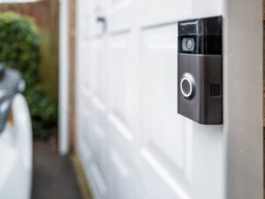 Close-up of a smart doorbell with a camera mounted on a white garage door, with part of a white car and greenery visible in the blurred background.