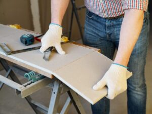 A person wearing white gloves uses a utility knife to cut a piece of drywall placed on a saw-horse workbench. A measuring tape and square are nearby, and the person is dressed in jeans and a checkered shirt.