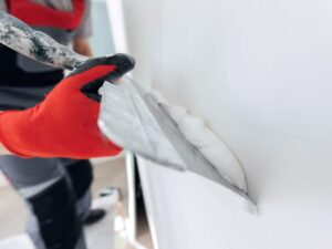 A person wearing a red glove uses a metal trowel to apply white plaster or joint compound to a wall, smoothing the surface during interior construction or renovation.