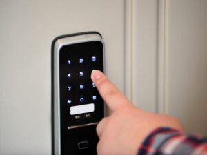 A person’s finger pressing a number on a digital keypad door lock, likely to enter a code and unlock the door. The keypad is illuminated and mounted on a white door.