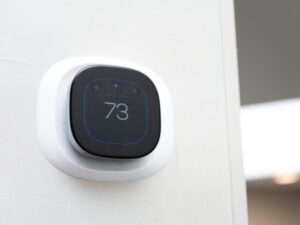 A modern digital thermostat mounted on a white wall displays the temperature set to 73 degrees. The device has a black screen with blue-lit numbers and icons, and a white frame.