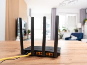 A black Wi-Fi router with four antennas and yellow cables connected sits on a wooden table in a bright, modern living room with blurred furniture and decor in the background.
