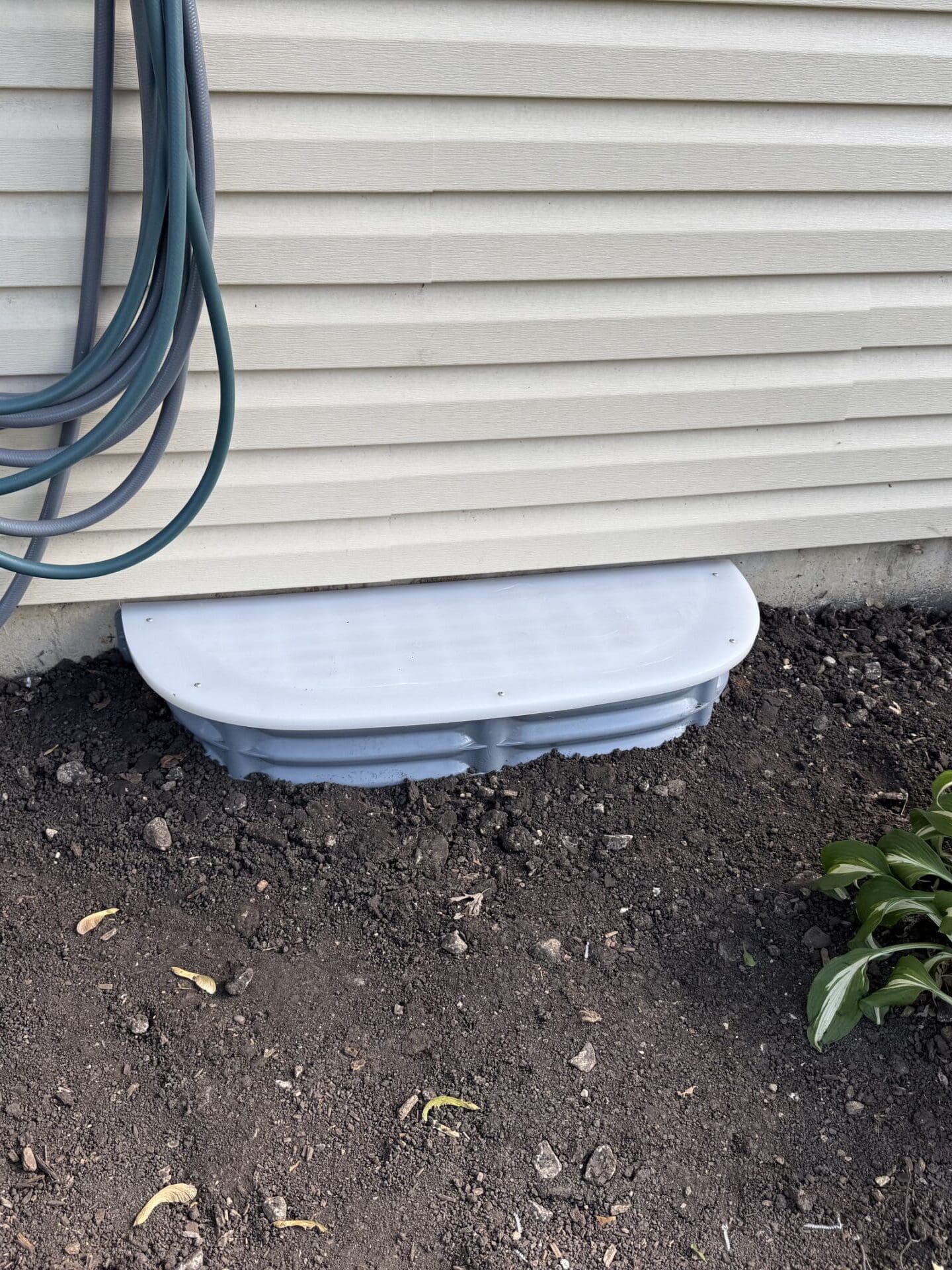 A white window well cover is installed against the beige siding of a house, with a coiled garden hose hanging nearby and green plants growing in freshly turned soil on both sides.