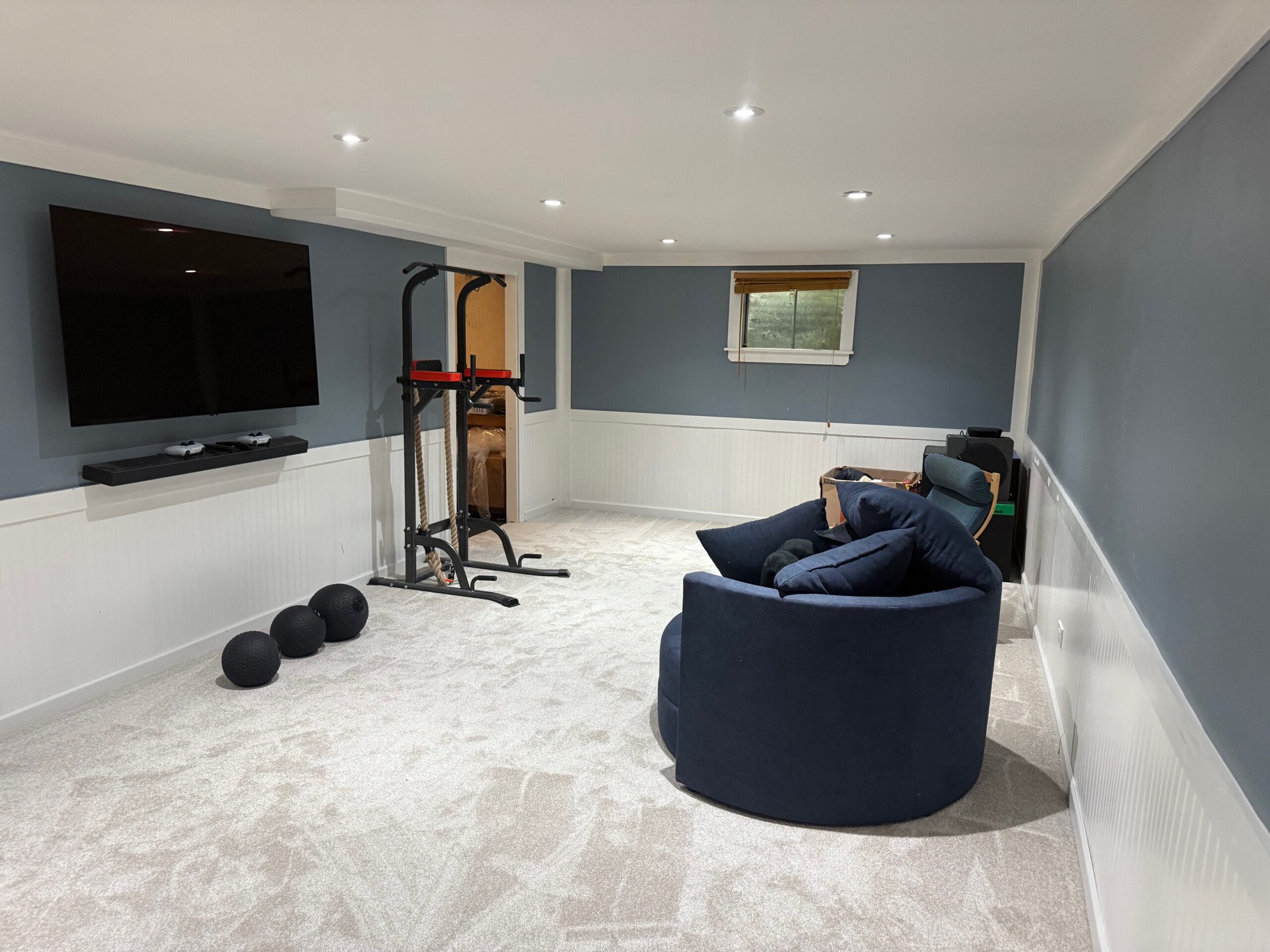 Brightwise - Basement Remodeling A basement room with grey-blue walls and carpet, featuring a large TV, an exercise station, three black medicine balls, two blue armchairs, and a small window letting in natural light.