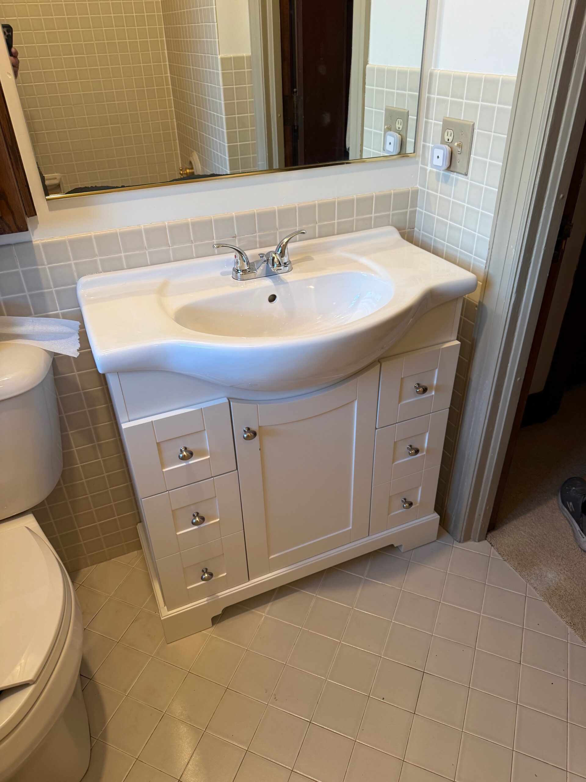 White bathroom vanity with a curved sink, chrome faucet, multiple small drawers, and a cabinet below, positioned against a tiled wall and beneath a large mirror. Part of a toilet and door are visible nearby.