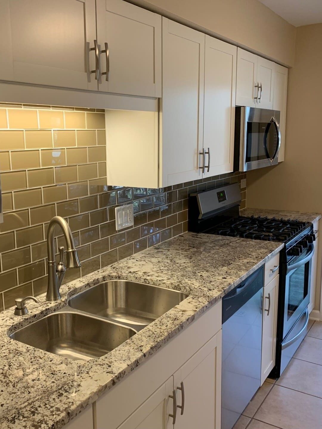 Modern kitchen with granite countertops, double sink, stainless steel appliances, white cabinets, beige tile backsplash, and large sliding glass door leading to a green outdoor area.