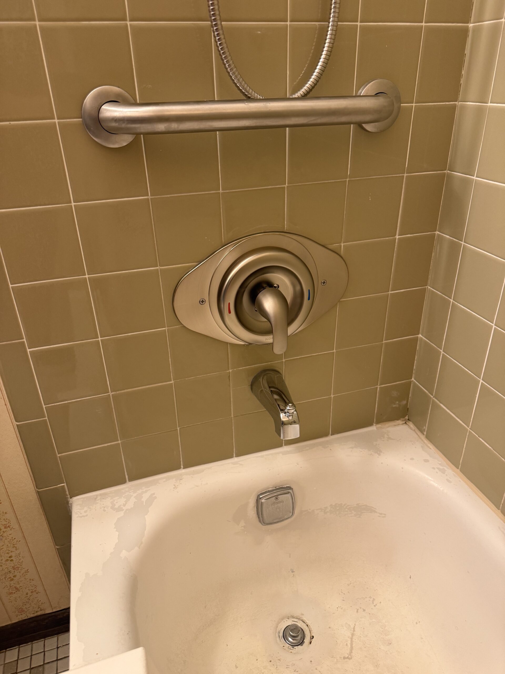 A beige tiled bathroom wall with a metal grab bar, a modern shower control lever, and a white bathtub with some stains and a silver faucet.