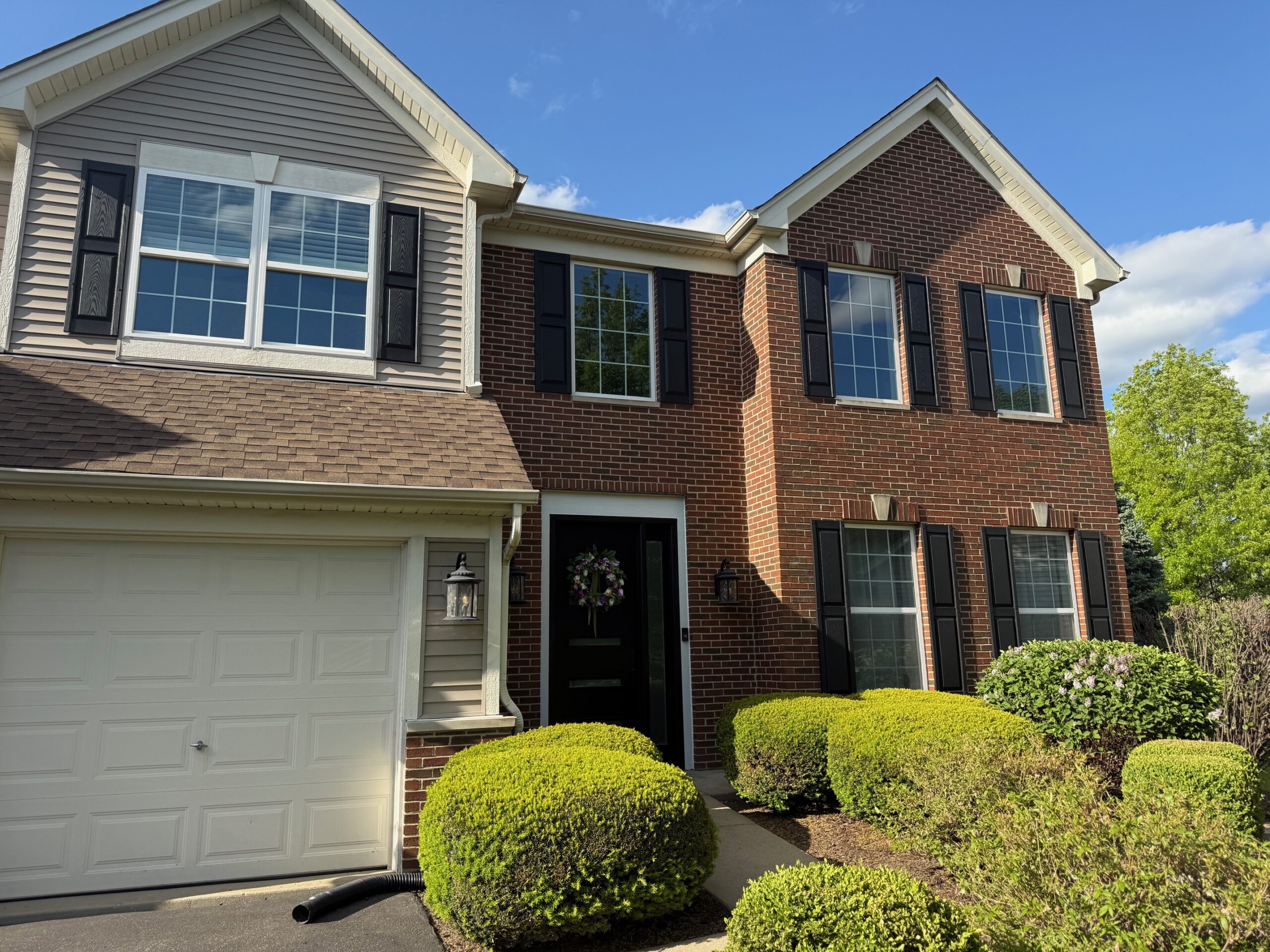 Two-story house with red brick and beige siding, black shutters, white-trimmed windows, a white garage door, and a dark front door with a wreath, surrounded by neatly trimmed green bushes under a blue sky.